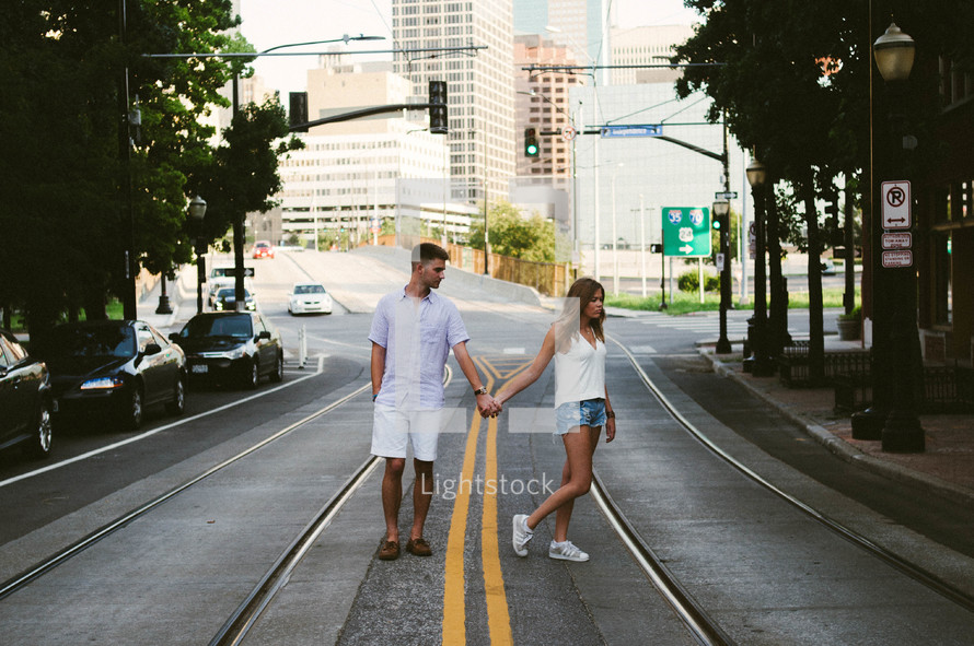 A young man and woman holding hands and walking together down a city street.