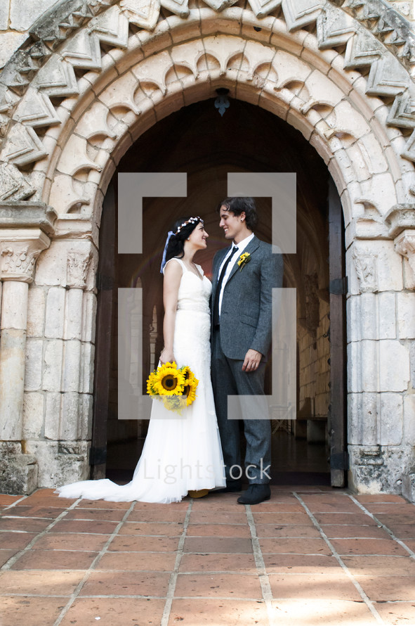 Bride with sunflower bouquet standing with groom on terra cotta tile in archway of church.