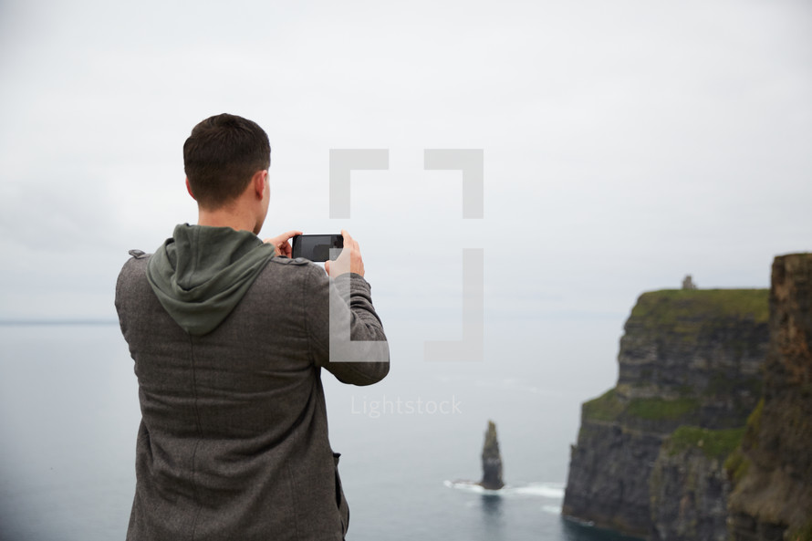 a man standing on the edge of a cliff taking a picture of the ocean 