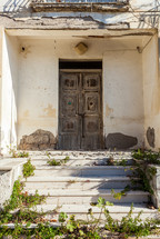 Old wooden door on the facade of an abandoned house with plants growing on the steps