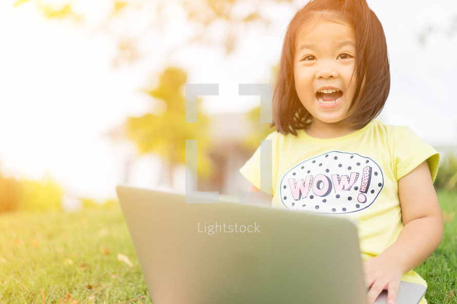 a toddler girl with a laptop in lap sitting in the grass