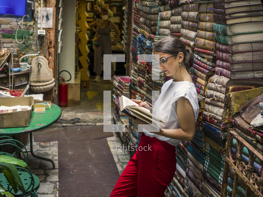 a woman reading a book near a stack of old books 