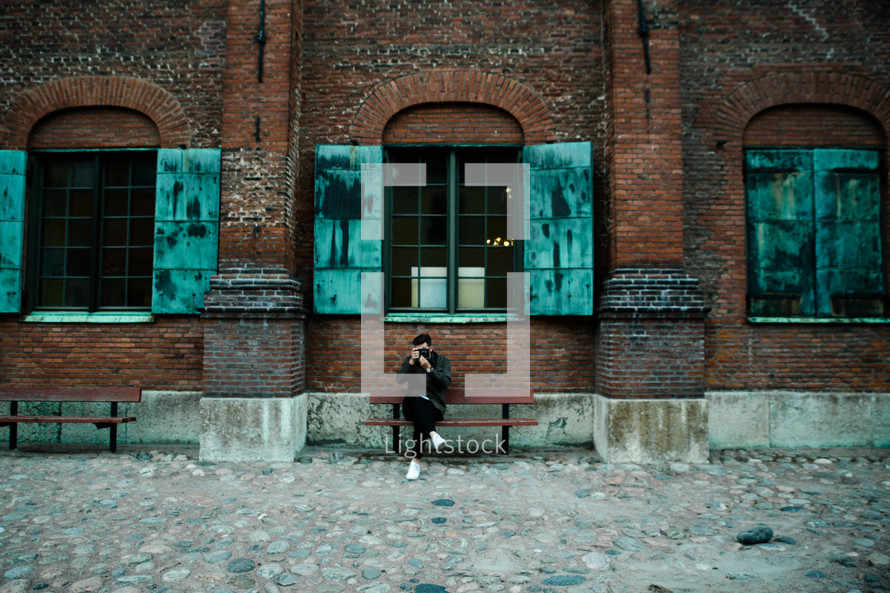 a man sitting on a bench in front of a warehouse taking a picture with a camera 