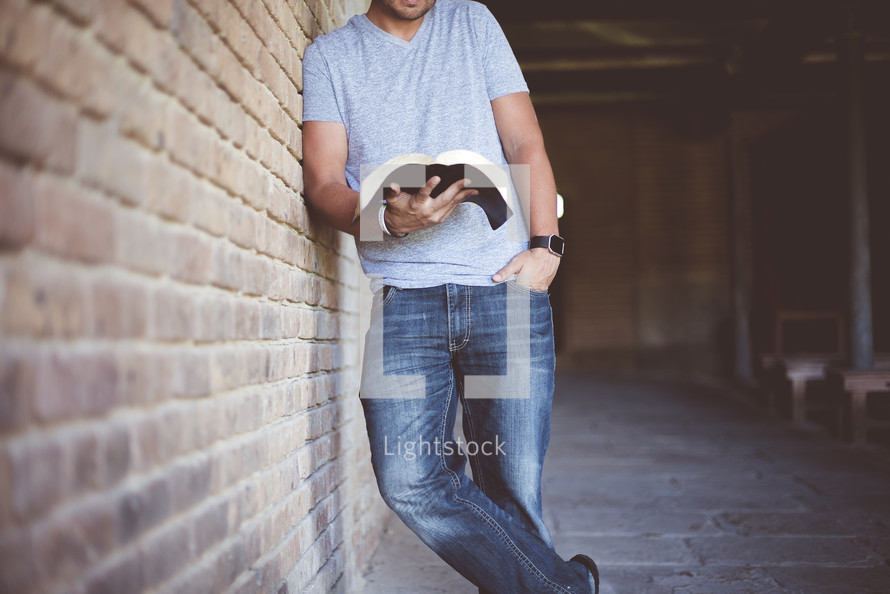 a man leaning against a brick wall reading a Bible 