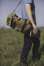 a man with a bag walking in a field of tall grass carrying a Bible 