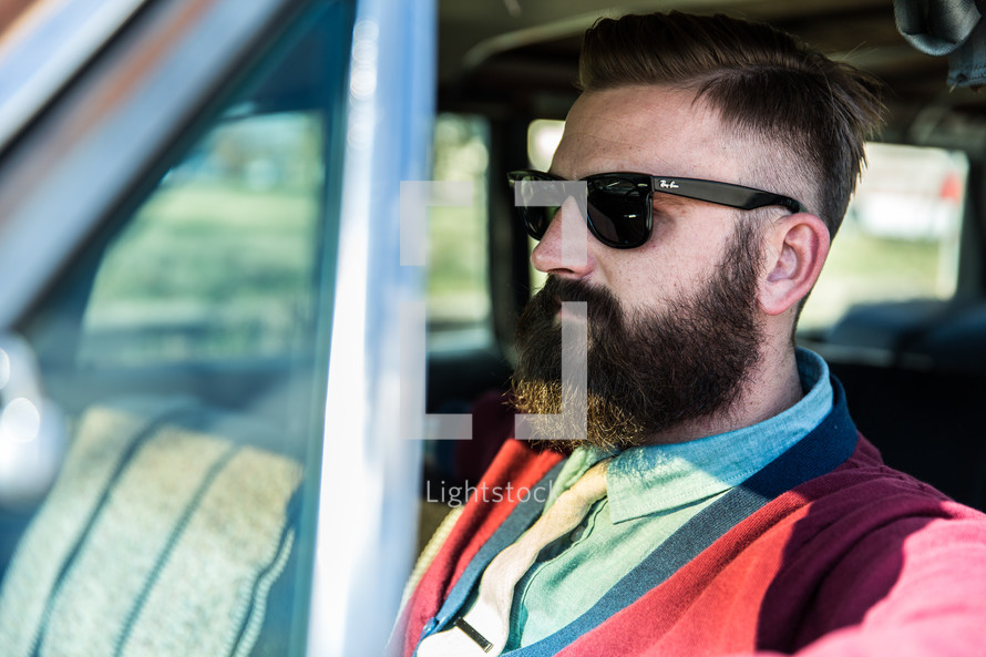 A man with a beard and sunglasses sitting in a car.