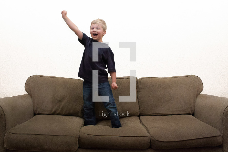 Boy standing on couch with hand raised in excitement with white background.