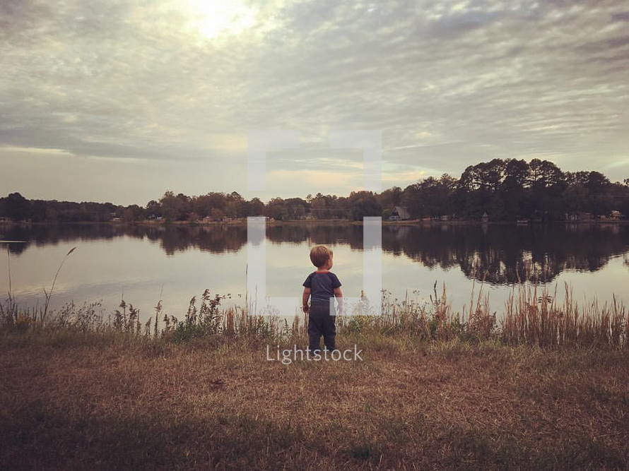 a toddler boy standing on a lake shore 