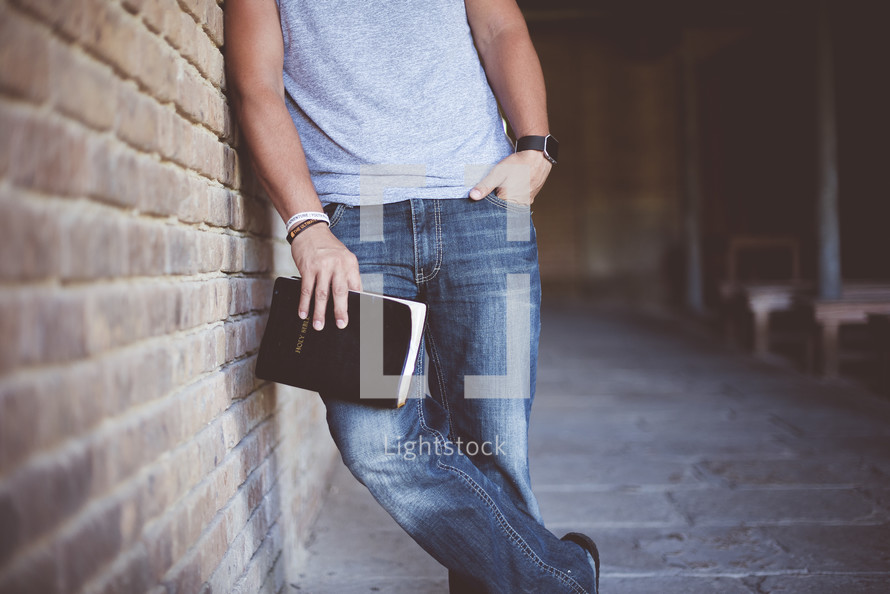 a man leaning against a brick wall holding a Bible 