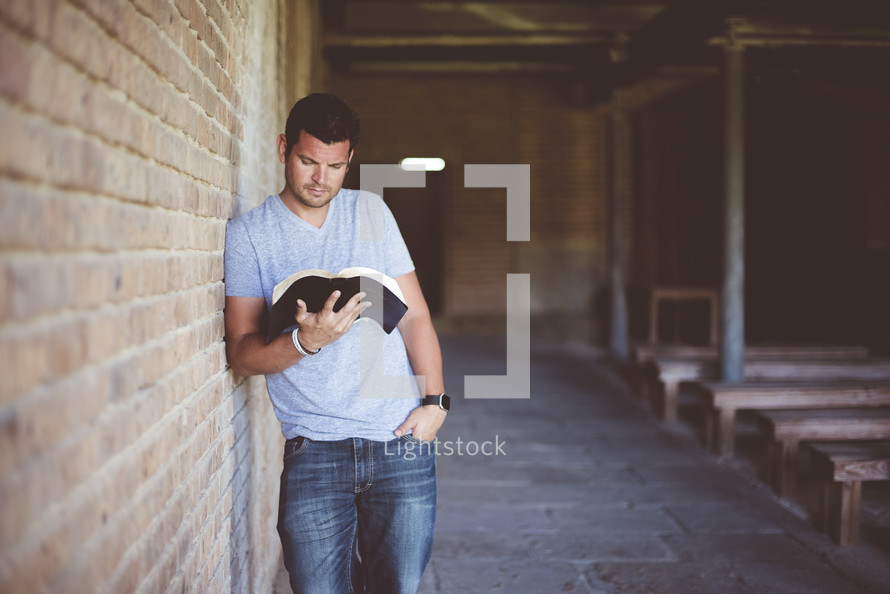 a man leaning against a brick wall reading a Bible 