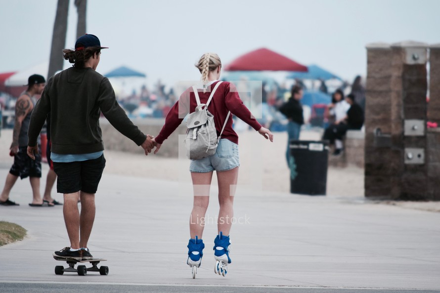 an adorable couple holding hands on a beach boardwalk while they roller blade and skateboard 