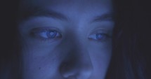 A young woman stares off camera with a soft blue light and static reflected on her face and in her eyes