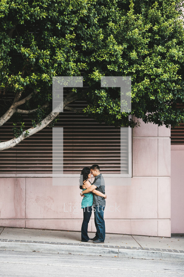 couple kissing under a tree standing on a sidewalk