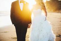 torso of a bride and groom holding hands walking on a beach