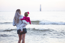 a woman standing by the ocean holding her toddler daughter 