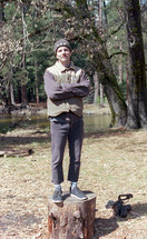 a man standing on a tree stump with arms folded across his chest 