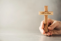 Hands holding wooden christian cross with text He is risen on grey background. 
