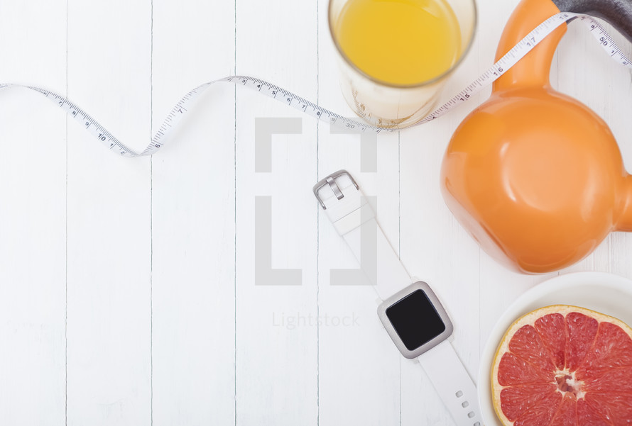 grapefruit, measuring tape, orange juice, kettle ball weight, and smartwatch fitness tracker on a white background 