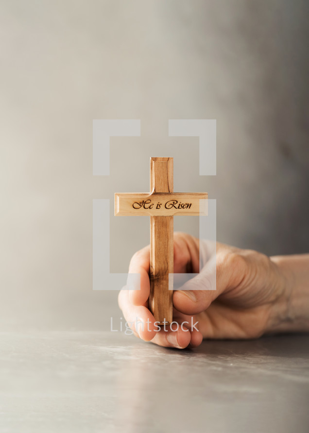 Hands holding wooden christian cross with text He is risen on grey background. 