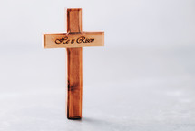 Wooden cross with text "He is risen" on grey background. 