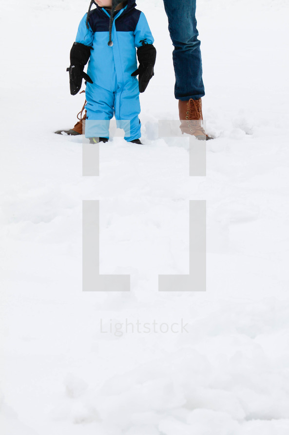 a toddler boy in a snowsuit standing in front of dad 