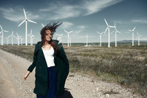 a woman walking on a dirt road beside of a wind farm 