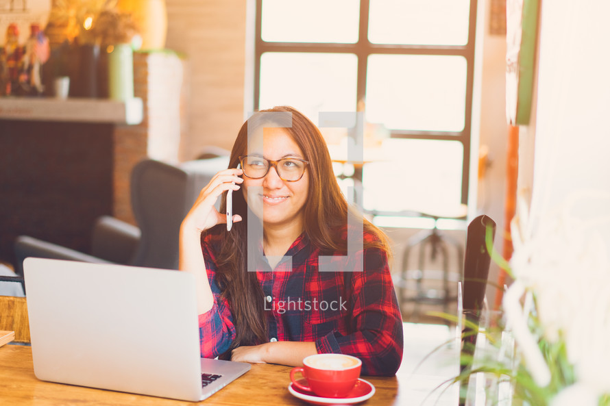 a woman talking on a phone sitting by a laptop with coffee 