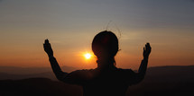 A woman praying with hands raised at sunset.