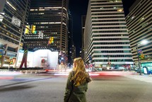 a woman standing looking up a skyscrapers in a city at night 