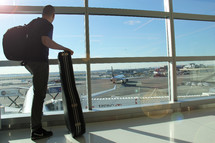 Man holding a guitar case looking out a window at an airport 
