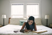A teen girl taking notes and studying the Bible on her bed.