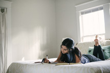 A teen girl taking notes and studying the Bible while laying on her bed.