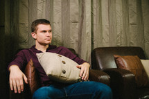 man sitting on leather chair against a wood wall, holding a pillow