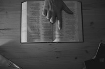 Hand pointing to scripture in a Bible on a wooden table.