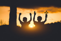 two boys with hands raised holding a cross against an orange sky at sunset 
