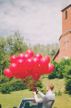 a man holding a bunch of red balloons in front of a church 
