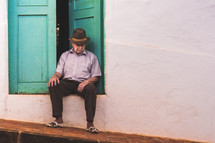 an elderly man sitting in a doorway looking down 