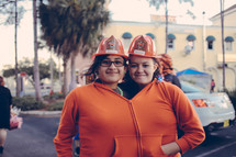 Two girls in hard hats zipped into one orange jacket.