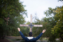 teen standing outdoors with raised hands near a wood cross 