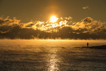 rising steam over ocean water and a man standing on a shore at sunrise 