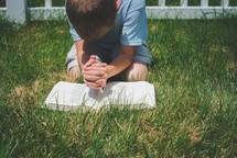 a boy kneeling in the grass reading a Bible and praying 
