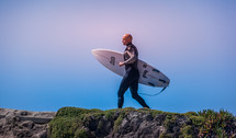 a surfer carrying his surfboard on a rocky shore 