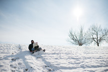 Father and child sledding down a hill in the snow.