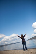 Woman standing at the water's edge with arms raised in praise.