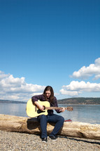 Smiling man sitting on beachwood at water's edge playing guitar.