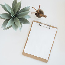 A clipboard with blank paper, cup of pencils, and a succulent plant on a white surface.