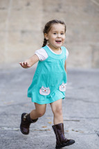 A little girl running in  a blue dress and boots.