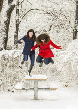Snowy trees with two girls jumping off a picnic bench covered with snow .