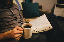 A man reading a Bible and holding a cup of coffee in his living room. 