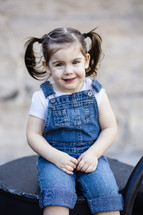 A little girl in overalls sitting down.